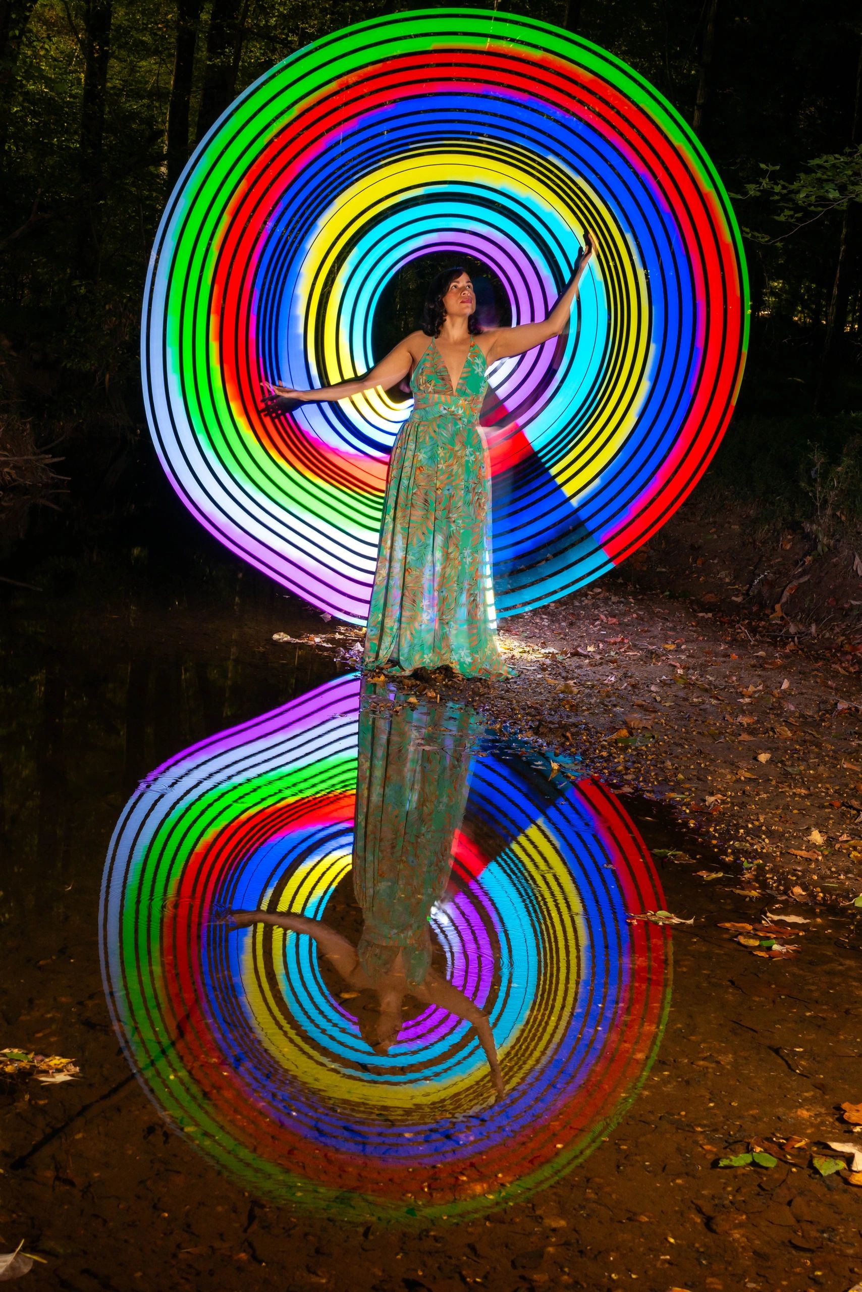 Woman in a floral dress stands before a vibrant rainbow light circle with reflective water below.
