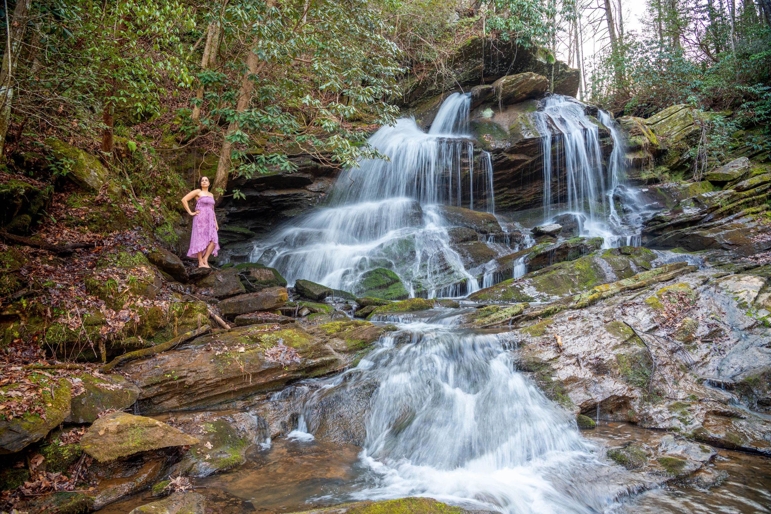 Woman in pink dress standing near a cascading waterfall surrounded by rocks and trees.