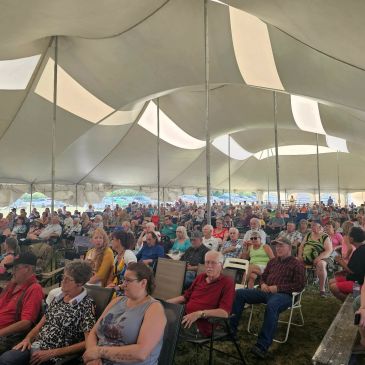 Large crowd seated under a spacious white tent at an outdoor event.