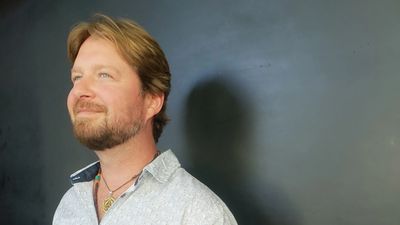 A man with light brown hair and a beard gazing thoughtfully to the side.