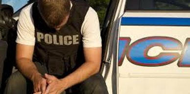 Police officer sitting with head down by police car, looking distressed.