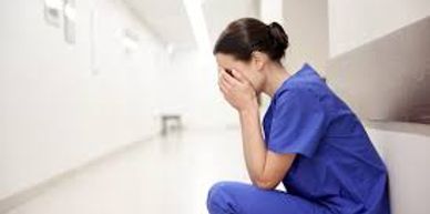 A nurse in blue scrubs sitting in a hallway, covering her face in distress.
