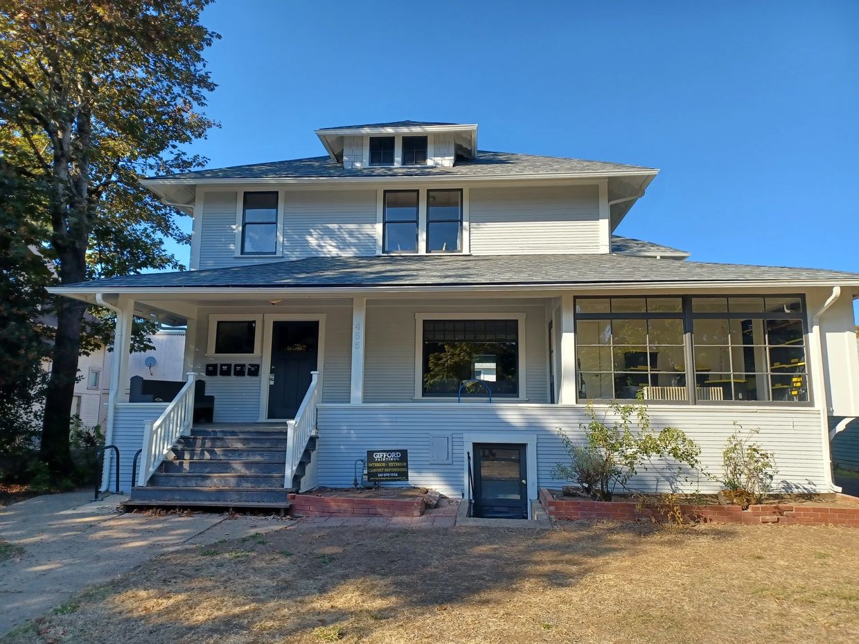A large white two-story house with a porch and basement entrance under clear blue skies.