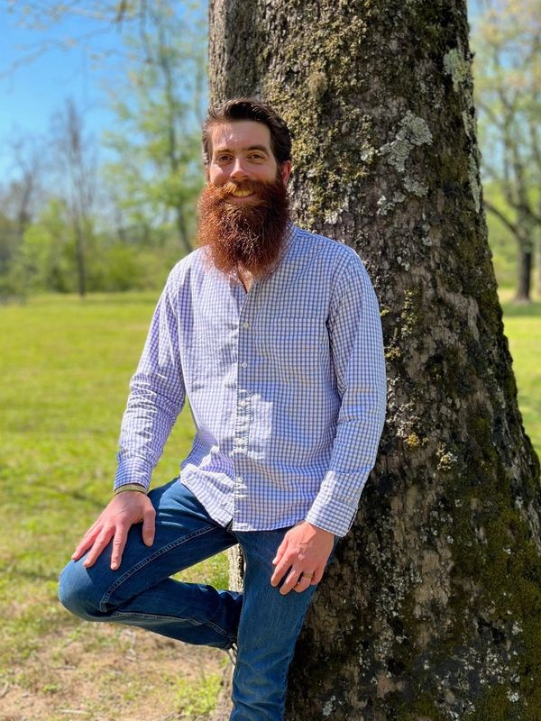 Man with a long beard leaning against a tree in a sunny park.