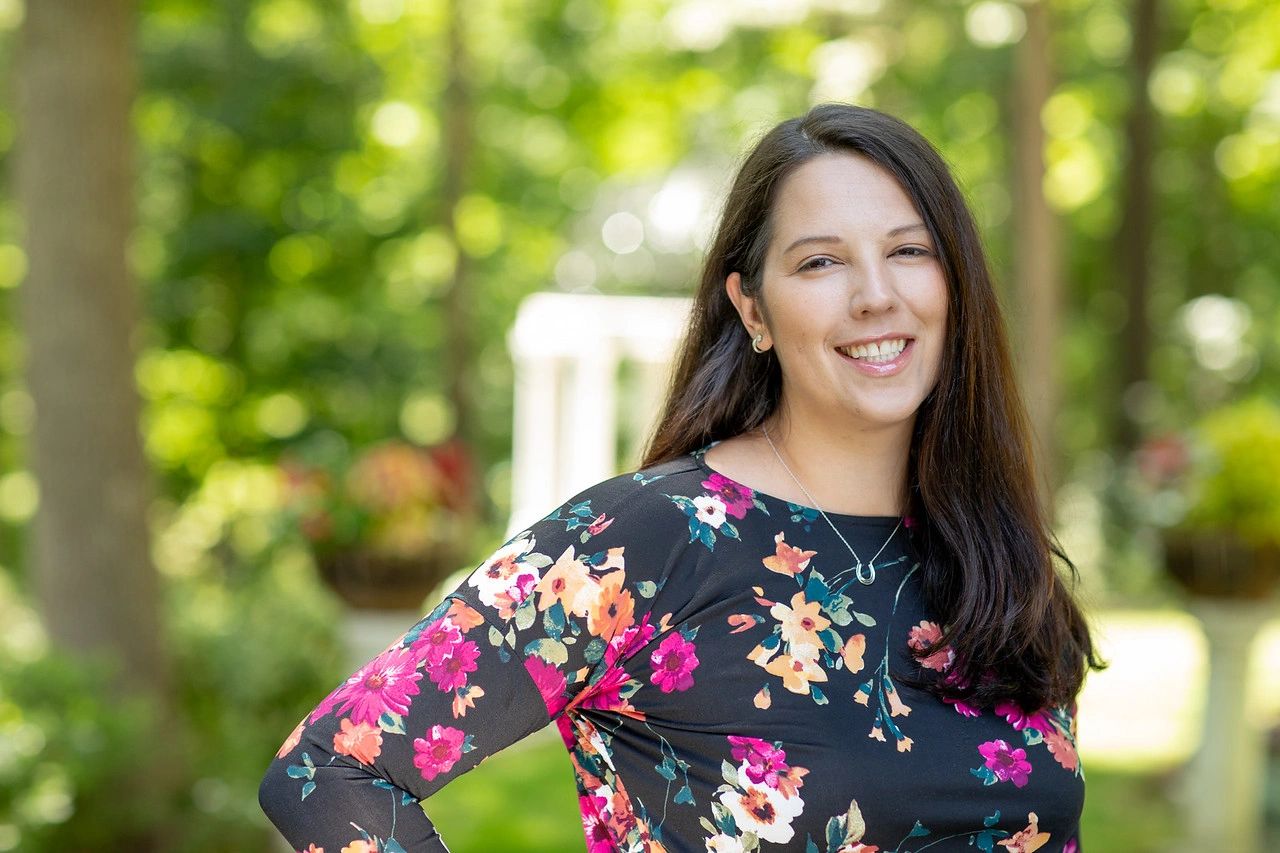 Smiling woman in a floral dress outdoors with greenery in the background.