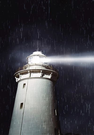 A lighthouse shines brightly in the rain against a dark night sky.