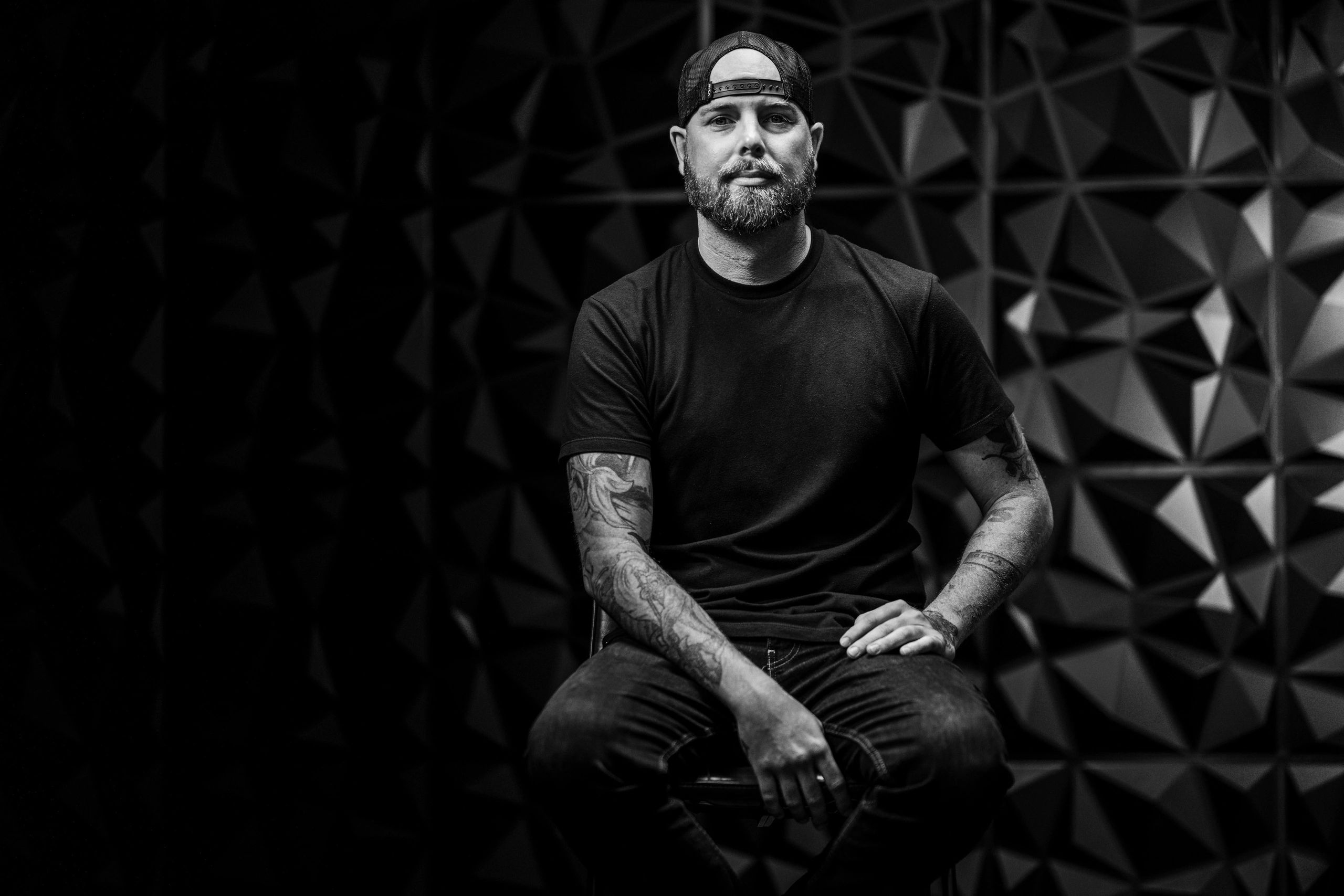 Bearded man with tattoos wearing a backward cap sitting against a geometric patterned wall.