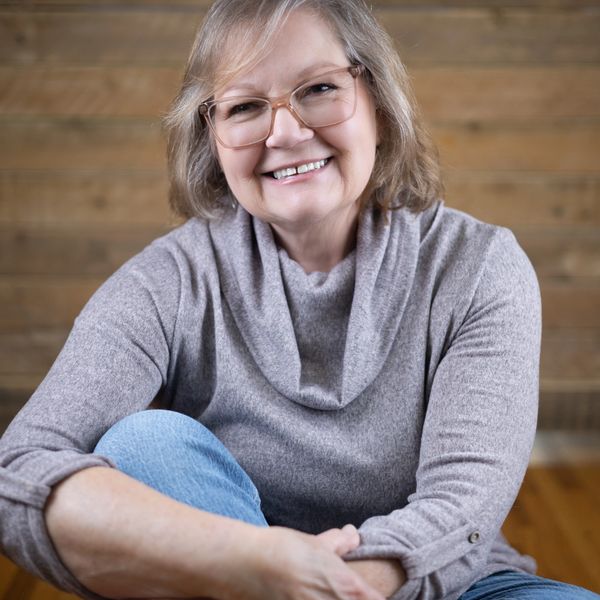 Smiling woman in glasses and cozy sweater sitting casually, with wooden background.