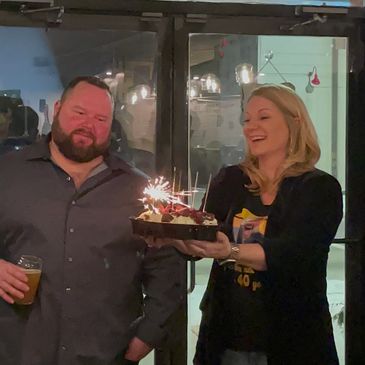 A woman presents a birthday cake with sparklers to a smiling man holding a drink.