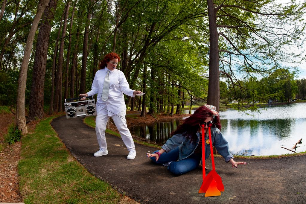 Two women on a path by a lake, one holding a boombox, the other with exaggerated red paper tears.
