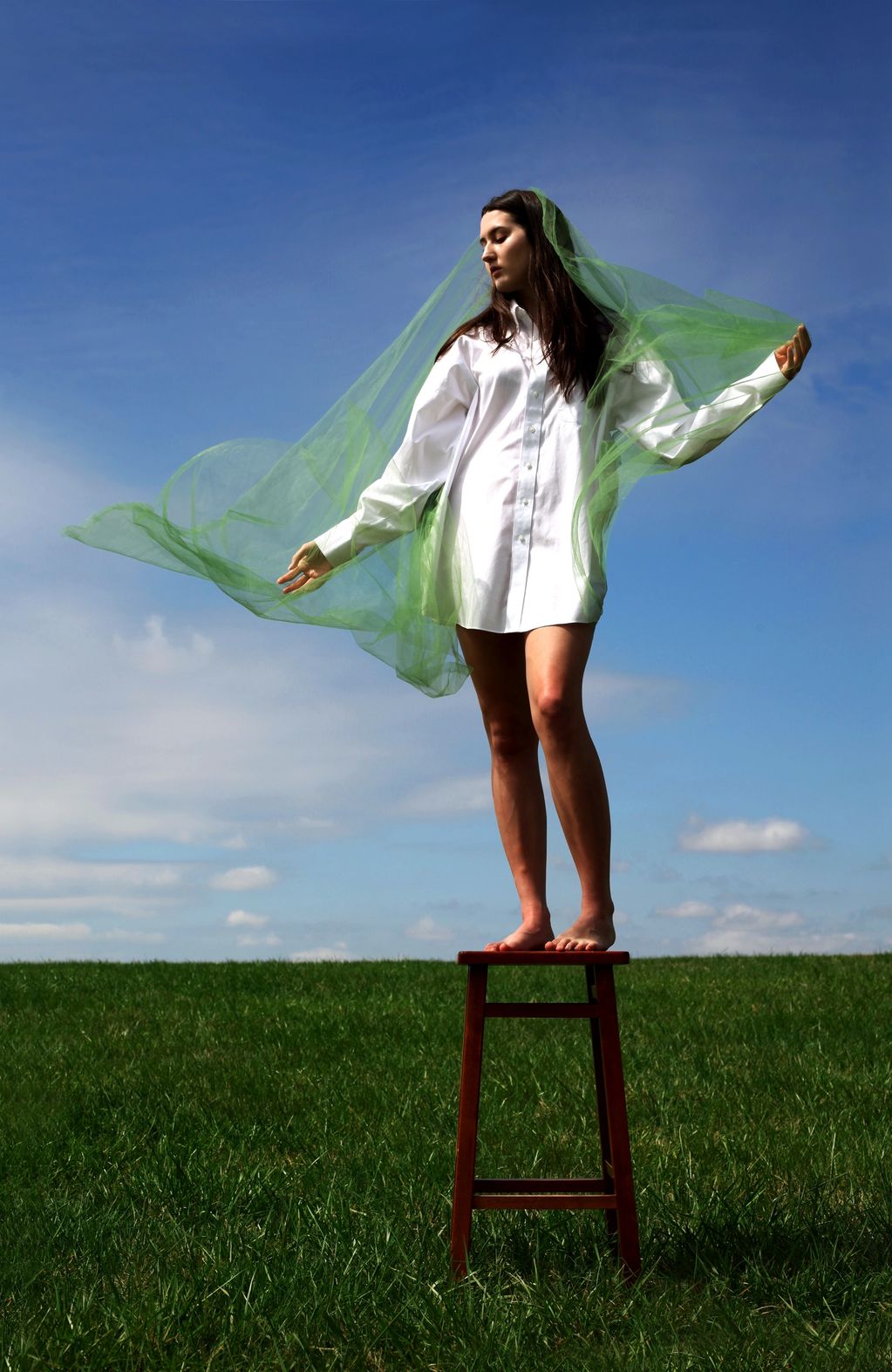 Woman in white shirt dress stands barefoot on stool with flowing green veil outdoors.