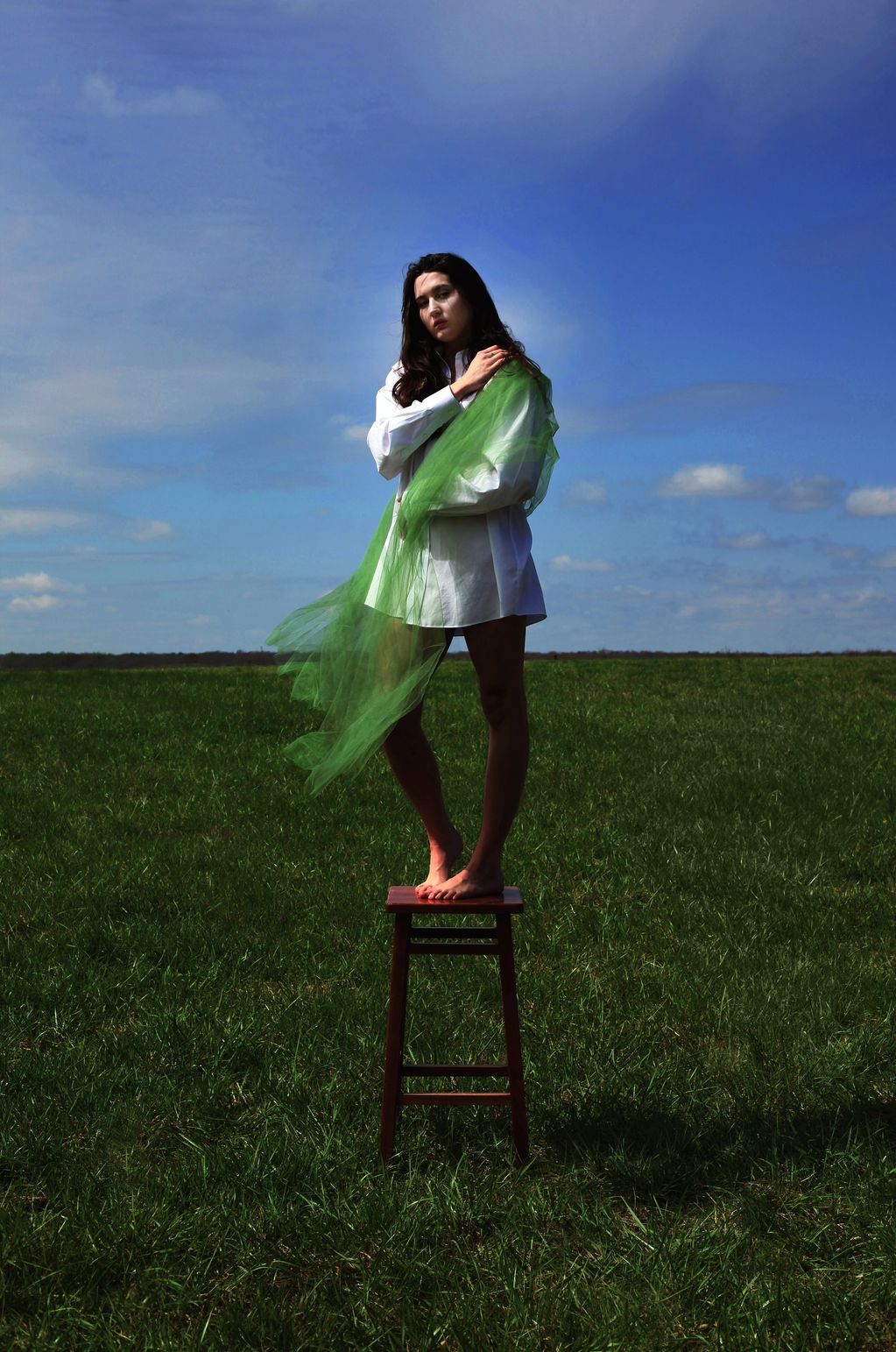 Woman in white shirt with green fabric stands barefoot on a stool in a grassy field.