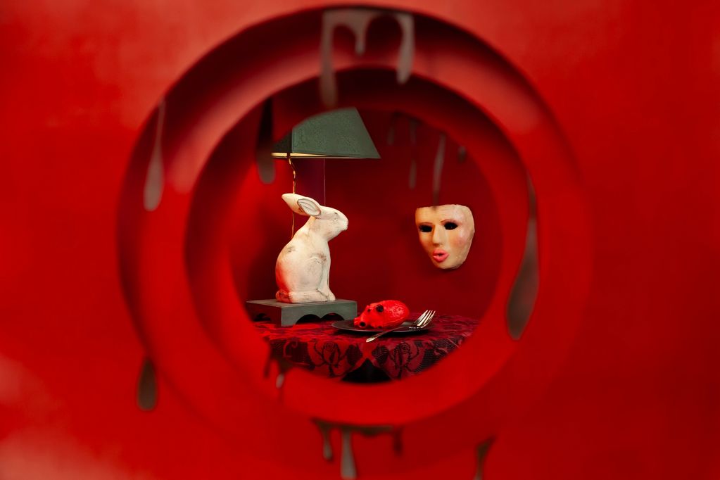 Surreal scene with a white rabbit sculpture, a floating mask, and a red mouse on a table inside a red circular frame.