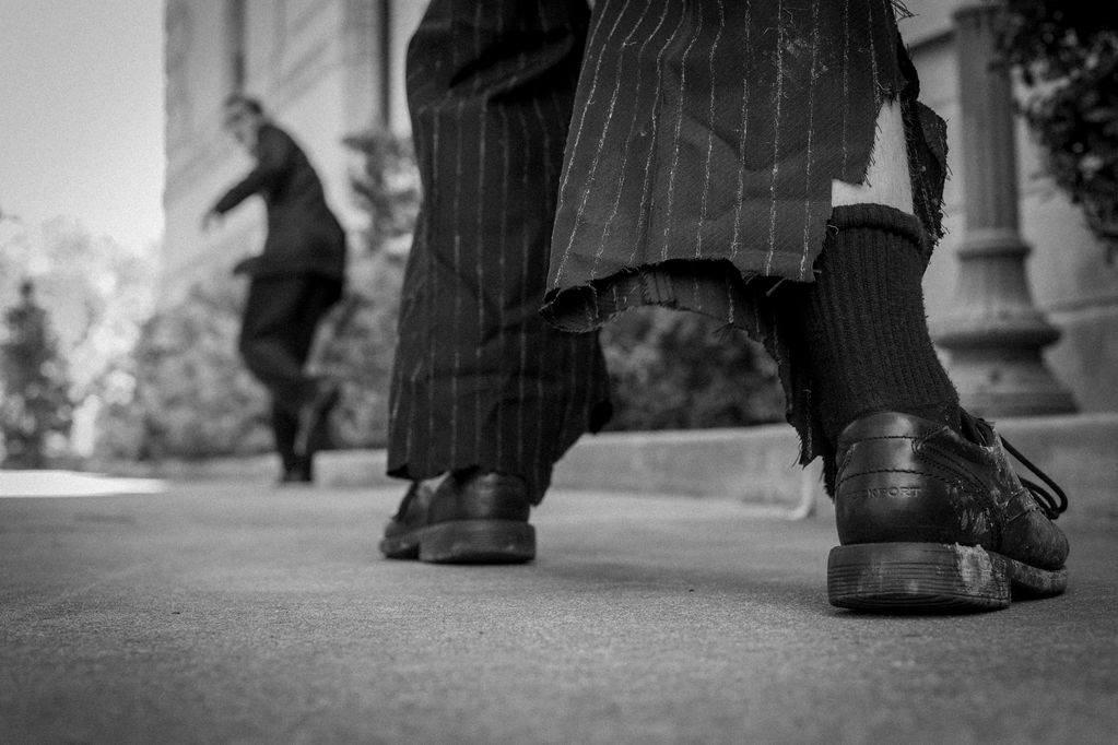 Close-up of worn shoes and frayed pants with a person running in the background.
