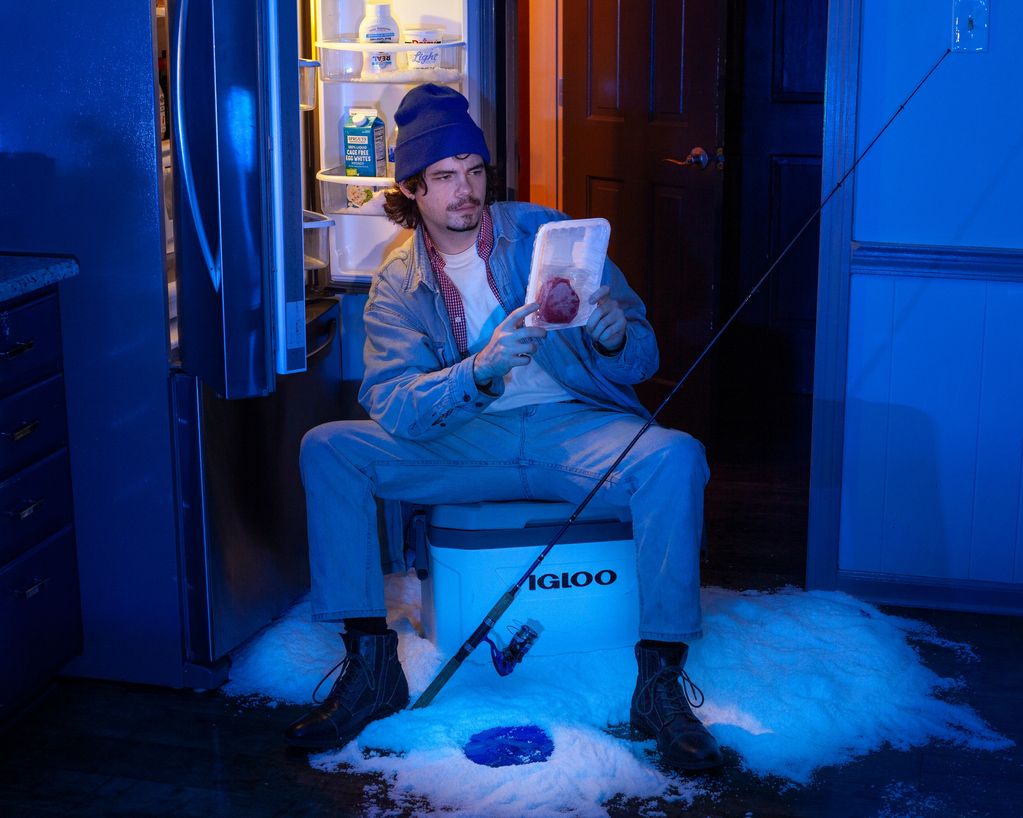 A man ice fishing indoors with a fishing rod and cooler near an open fridge.