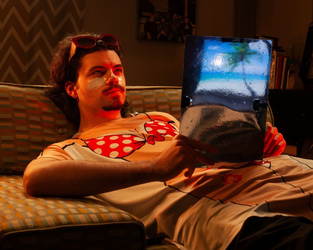 Man lounges on couch with sunscreen and sunglasses, holding a reflective picture of a beach scene.
