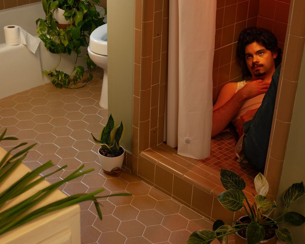 A man sits curled up in a tiled shower stall surrounded by plants in a bathroom.