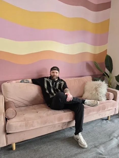 Man sitting on a pink sofa with a colorful wavy wall behind.