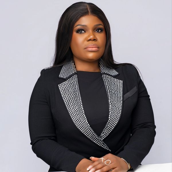 Confident woman in a black blazer with decorative collar, posing against a plain background.
