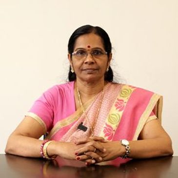 Middle-aged woman in traditional pink saree sitting with folded hands.