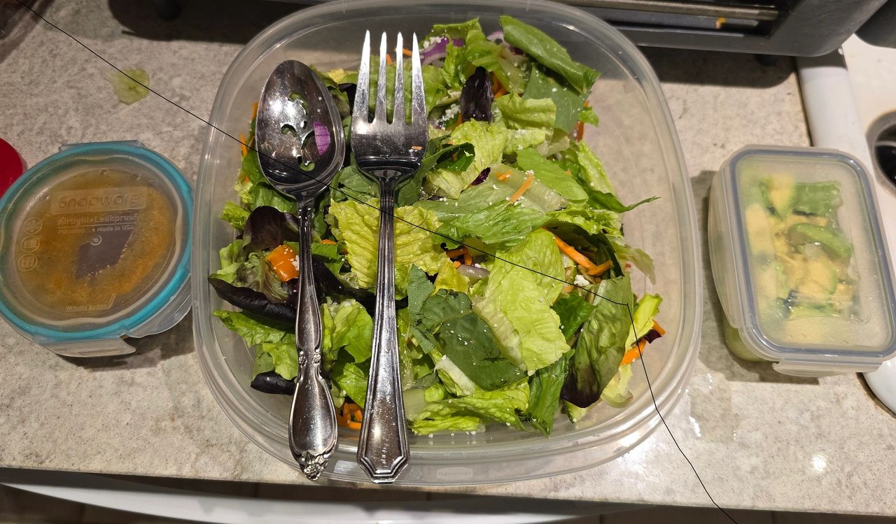 Containers of prepared salad ingredients for Grilled Nectarine & Chicken Salad on a kitchen counter. The image shows a large plastic container of mixed greens with silver serving utensils, a small container of diced avocado, and a jar of honey-chile-lime vinaigrette.