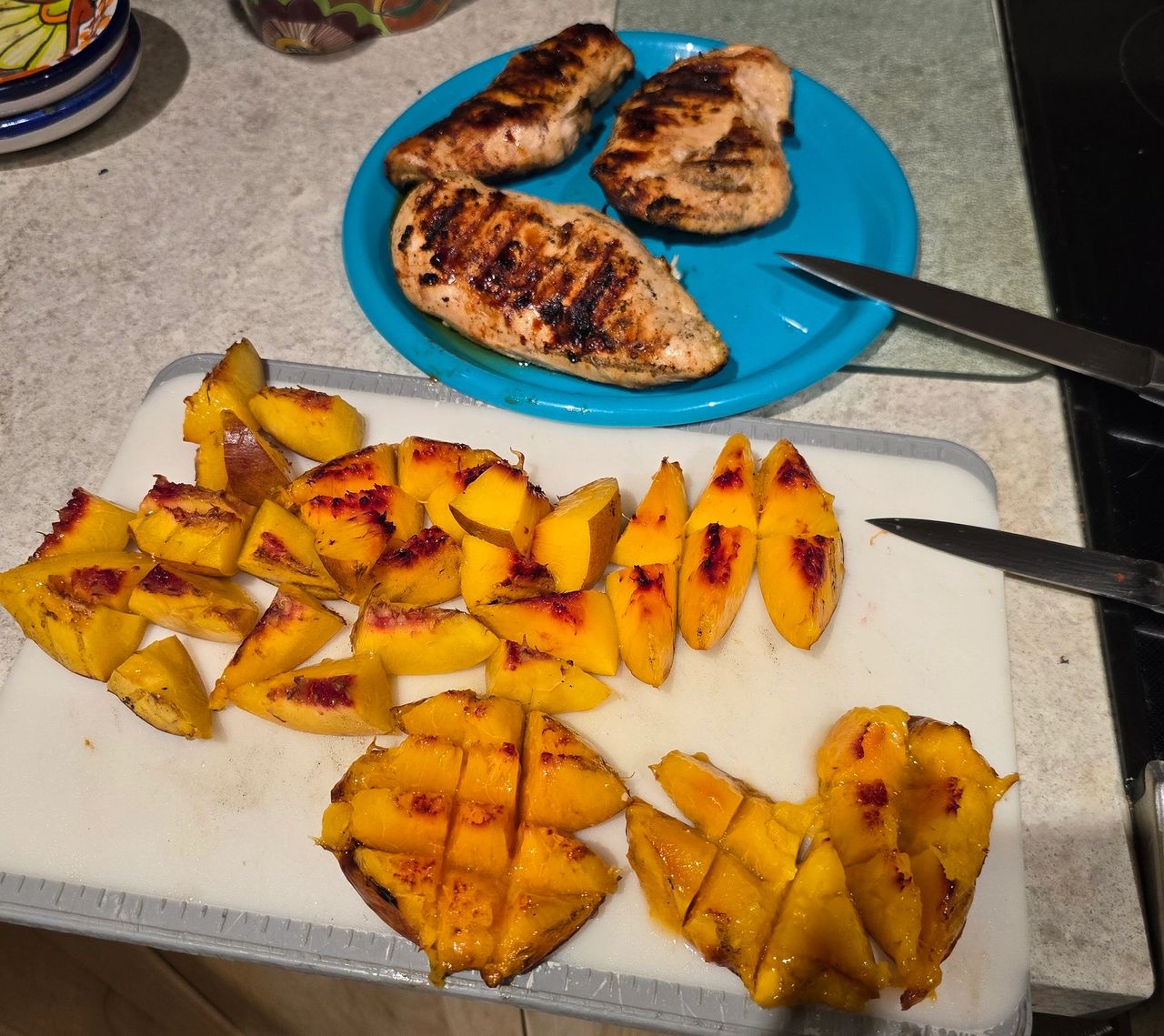 Grilled chicken breasts resting on a blue plate beside a cutting board filled with sliced, charred nectarines. The counter also includes a knife and grilled fruit pieces arranged in bite-sized portions, ready for salad assembly.