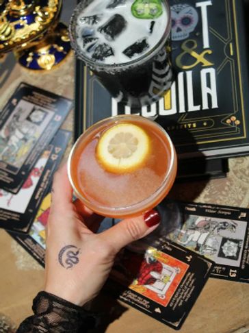 Two cocktails on a table with tarot cards and a book about gin and tequila.
