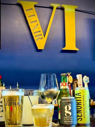 Assortment of drinks with bold yellow 'ELIXIR VI' sign on blue wall.