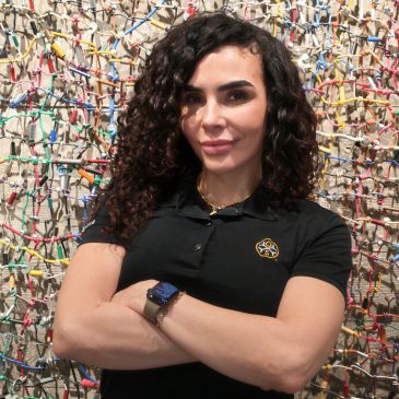 Confident woman with curly hair and black polo shirt stands in front of colorful wall art.