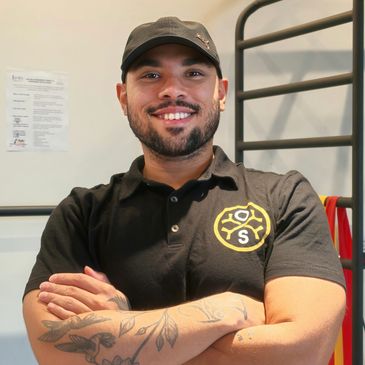Smiling man with tattoos in a black polo and cap.