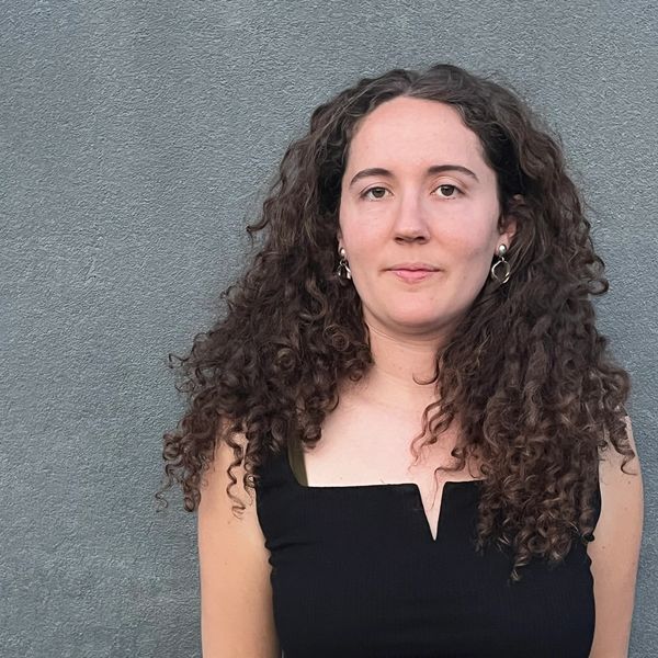 Young woman with curly hair stands against a gray wall.