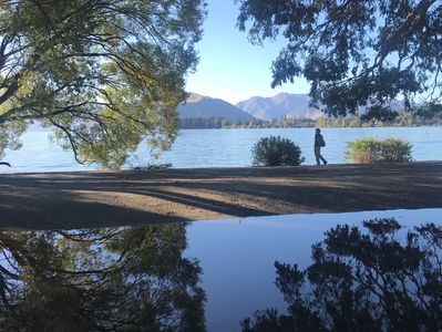 Person walking by a lake with trees and mountains in the background.