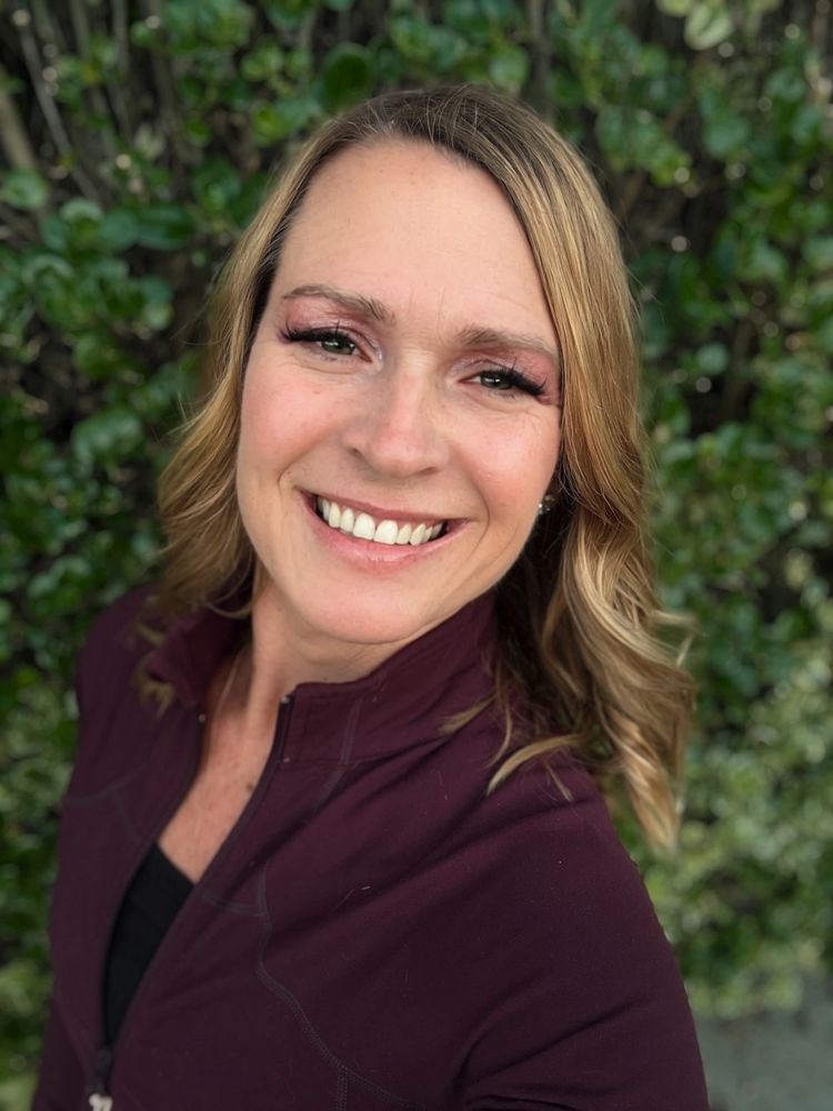 Smiling woman with blonde hair outdoors in a maroon jacket.