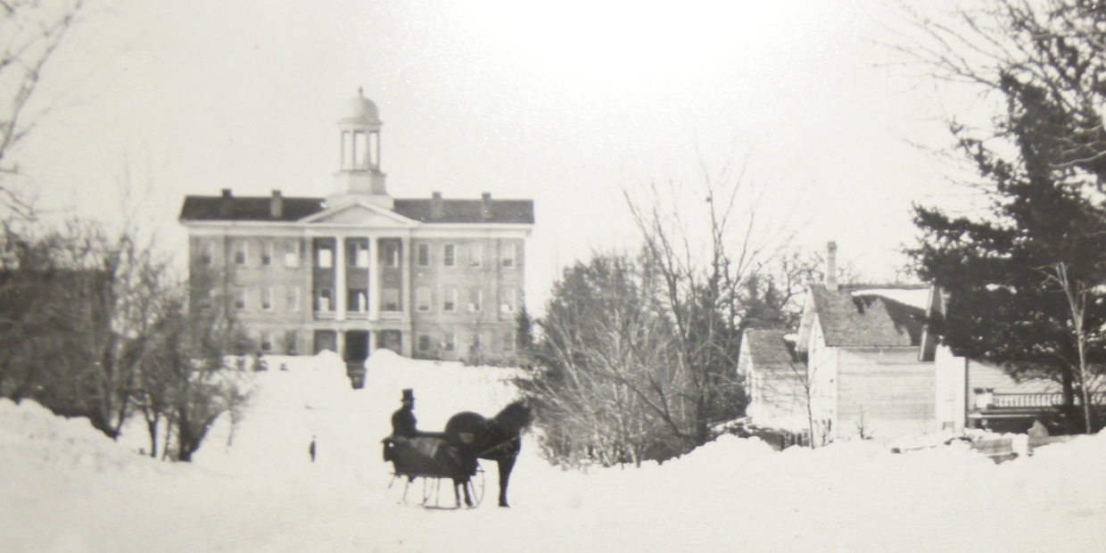 Early photo of Old Main in the winter with a horse and carriage in front on the snowy street.