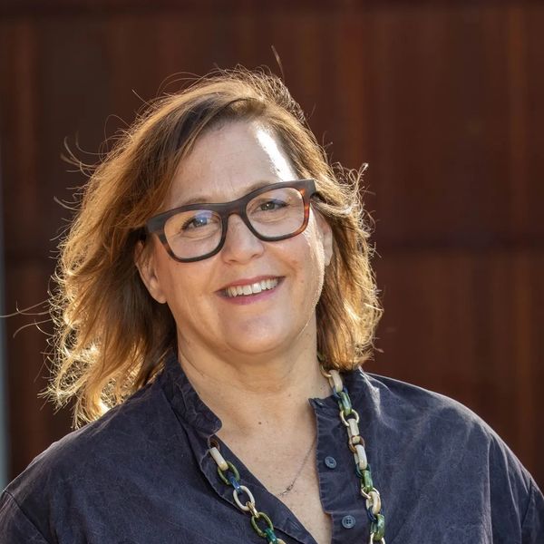 A middle aged woman with glasses, brown hair, a blue shirt and neckless standing outside. 