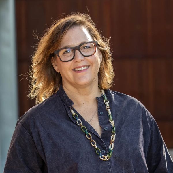 A middle aged woman with glasses, brown hair, a blue shirt and neckless standing outside. 