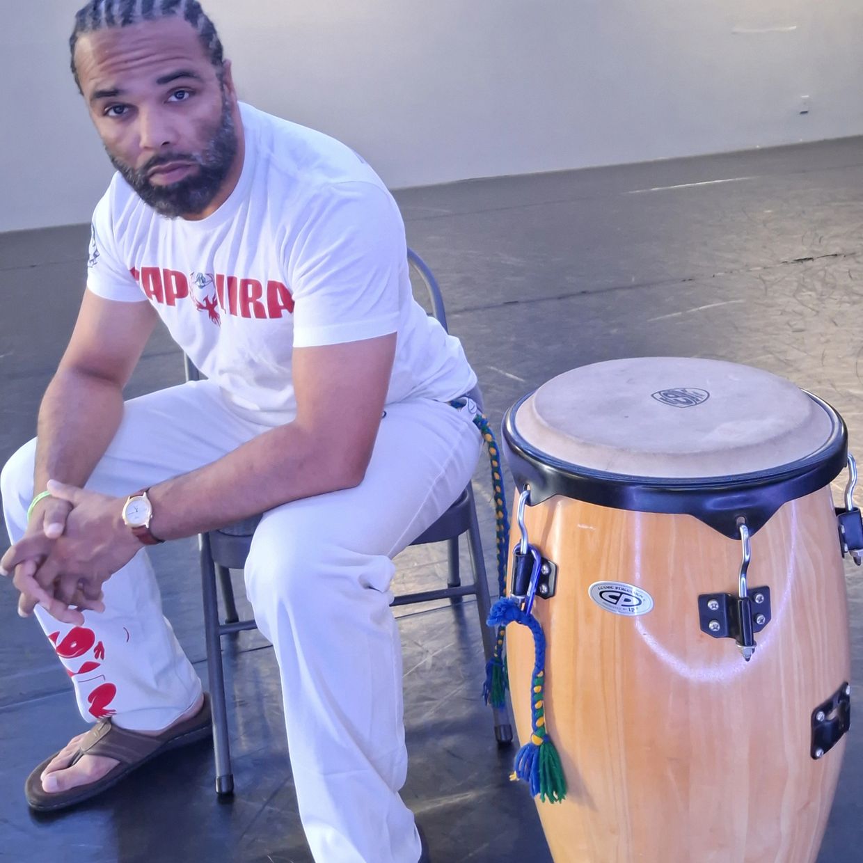 Man with braided hair sitting on a chair beside a conga drum in a spacious room.