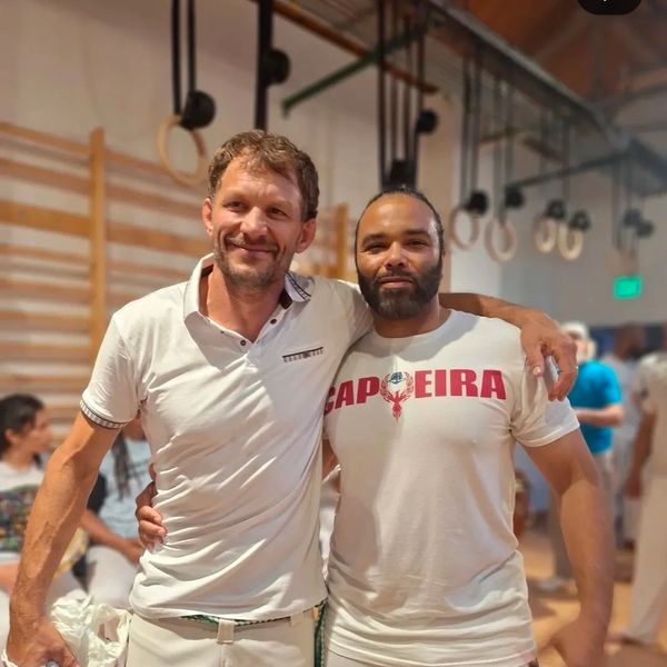 Two men smiling and posing together in a gym with gymnastic rings in the background.