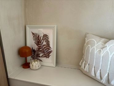 Cozy corner with a framed fern print, orange lamp, and textured cushion.