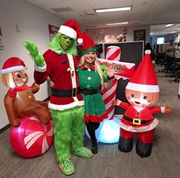 People dressed as the Grinch and an elf posing with inflatable Christmas decorations in an office.