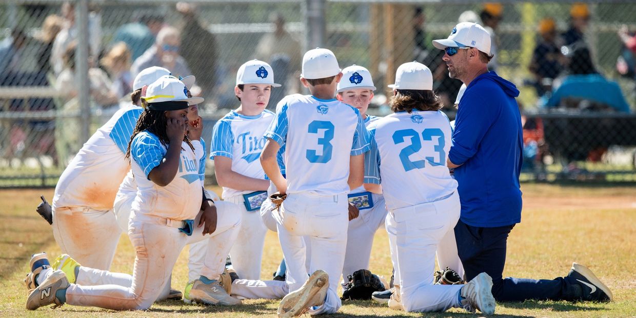 Youth baseball team in white and blue uniforms listens to coach on field.