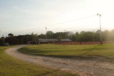A dirt path curves around a grassy field near buildings and trees at sunset.