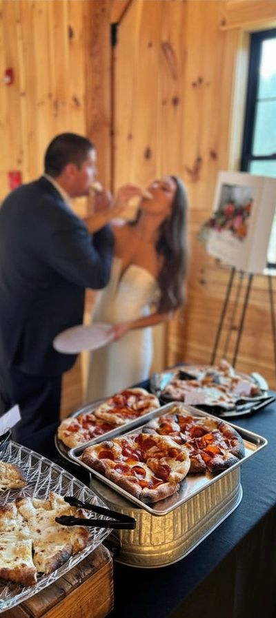 Newlyweds feeding each other pizza slices at a rustic wedding reception.