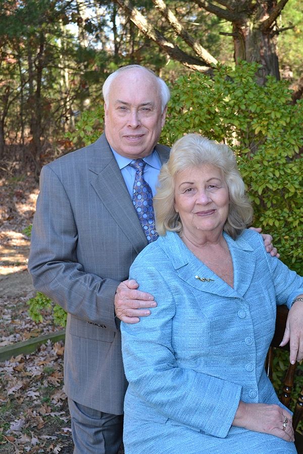 An elderly couple posing outdoors with warm smiles, dressed formally.