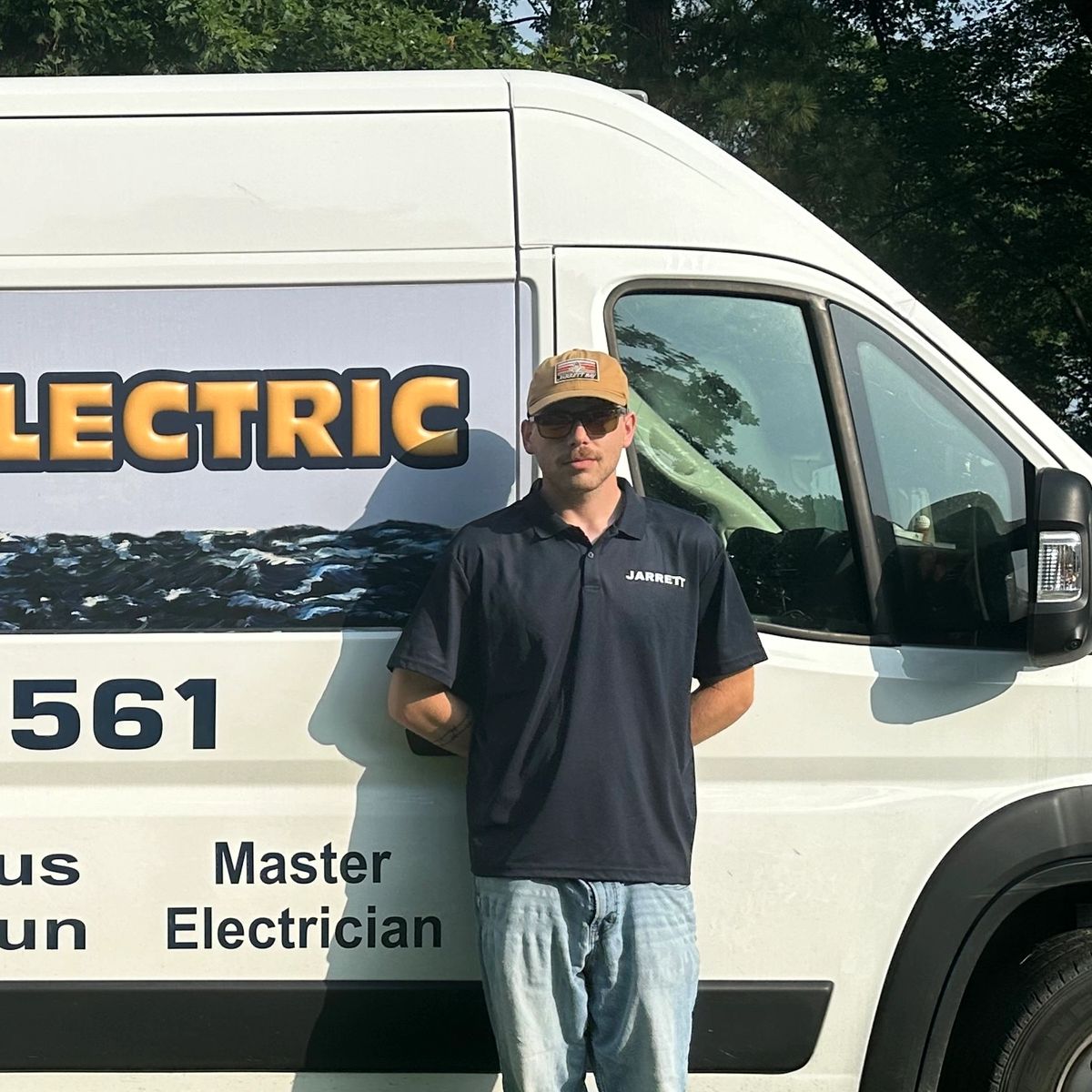 Man wearing sunglasses and cap stands by an electrician van.