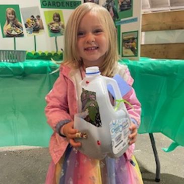 A young girl holding a milk jug with ladybugs, smiling at a gardening event.