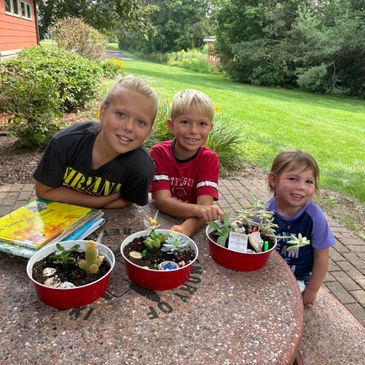 Three children proudly show their succulent plant pots outdoors.