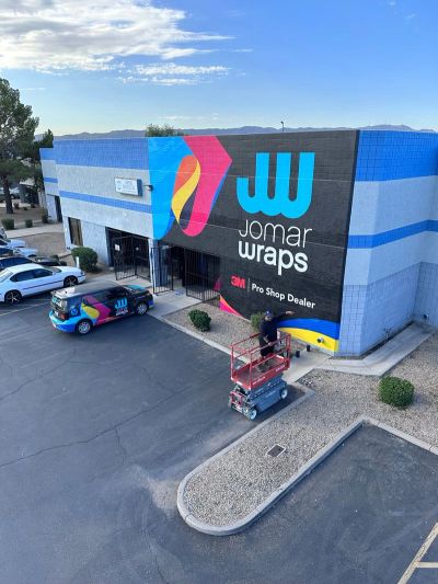 A vibrant building with Jomar Wraps branding and a worker on a lift painting the exterior.