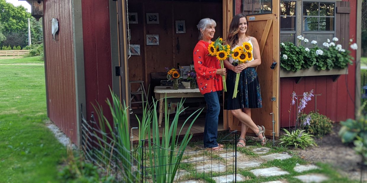 Two women holding sunflowers stand by a red garden shed.
