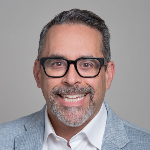 Smiling man wearing glasses and a light blue blazer against a gray background.